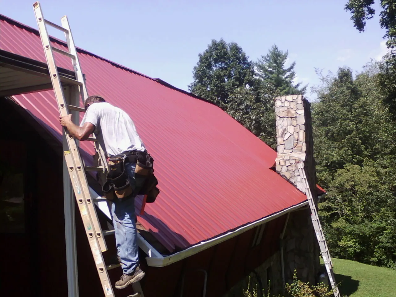 Roofer on a ladder beside a red metal roof and stone chimney — Soffit & Fascia Repair by Roofing Brothers in Evansville